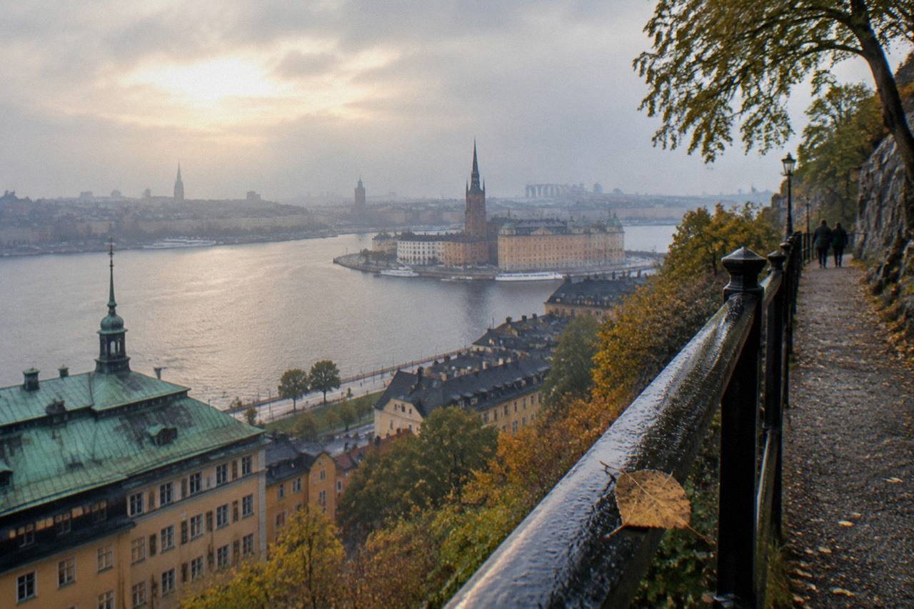 Evening light over Stockholm, Sweden, seen from the Monteliusvägen railing on Sodermalm, calm water below, copper spires and Gamla Stan rooftops across the frame, a walker pausing at the rail.
